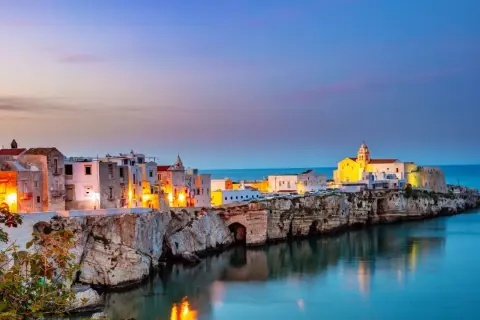 Vieste centro storico di sera, con case bianche illuminate e il mare blu che riflette le luci.