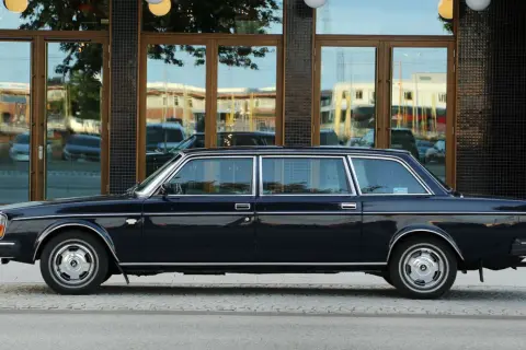 En mörkblå Volvo 264 TE, en lyxig limousin, står parkerad utanför en byggnad med stora fönster.