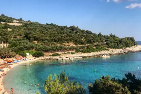 Słoneczna plaża na wyspie Vis, z turkusową wodą, zielonymi wzgórzami i ludźmi odpoczywającymi na piasku.