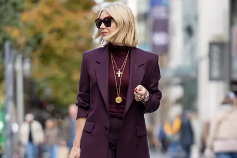 Elegancka kobieta w garniturze w kolorze burgundowym, z złotym krzyżykiem na szyi. Ten kolor pasuje do każdej pewnej siebie kobiety.