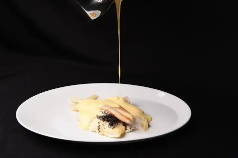 Ein Koch übergießt Fisch mit Spargel und Kaviar mit einer cremigen Sauce. Die Sauce sieht aus, als wäre die Sahne geronnen, was dem Gericht eine interessante Textur verleiht.
