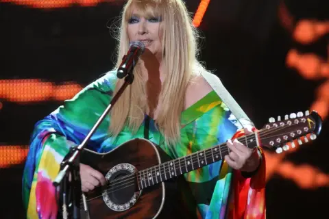 Maryla Rodowicz, ile ma lat, w kolorowym stroju gra na gitarze dwunastostrunowej na scenie.