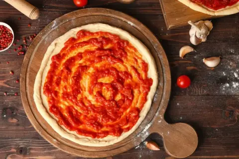 Une pâte à pizza crue est étalée sur une planche en bois, recouverte d'une sauce tomate maison. La question quelle sauce tomate pour pizza est au cœur de cette préparation.