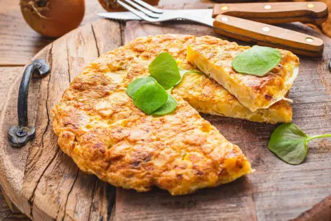 Goldbraune Tortilla de Patatas, garniert mit frischen Blättern, auf rustikalem Holzbrett. Dazu Zwiebeln und Besteck.