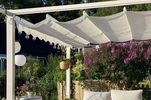 En vit kaprifol pergola med hängande ljusslingor och en frodig syrénbuske i bakgrunden.
