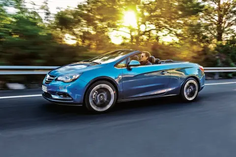 Niebieski Opel Cascada z otwartym dachem sunie po drodze, a słońce przebija się przez drzewa.