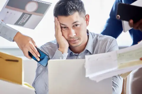 Zmęczony mężczyzna przy laptopie, otoczony dokumentami i telefonami. Czy to właśnie multitasking co to znaczy?
