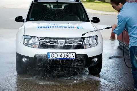 Mężczyzna myje białe Dacia Duster na myjni bezdotykowej, spryskując przód auta wodą pod ciśnieniem.