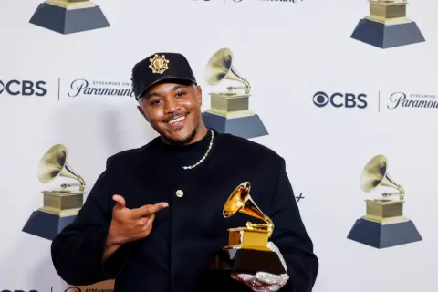 Khris Riddick-Tynes smiles, holding a Grammy award and pointing to it. He wears a black cap and jacket.