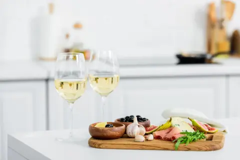 Due calici di vino bianco, ideali come vino bianco per cucinare, accompagnano un tagliere con formaggio, fichi, salumi e olive.