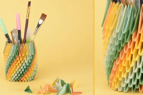 Bunter Papierkorb gefüllt mit Stiften und Pinseln, daneben Papierbögen und kleine Origami-Figuren.