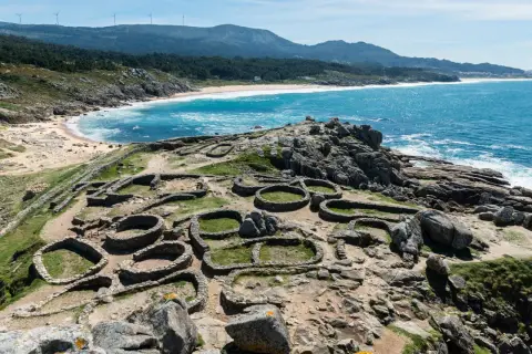 Ruinas circulares de un castro gallego junto a una playa de aguas turquesas. Paisajes de Galicia que evocan historia y naturaleza.