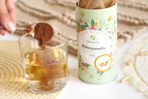 Łyżeczka z proszkiem ashwagandhy wsypywana do szklanki wody. Opakowanie Naturalis Ashwagandha BIO obok.