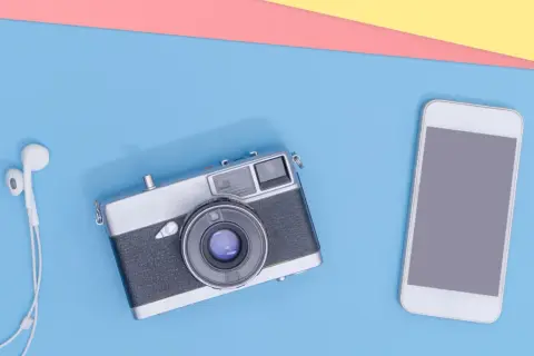 Un smartphone et un appareil photo argentique vintage côte à côte, avec des écouteurs blancs.