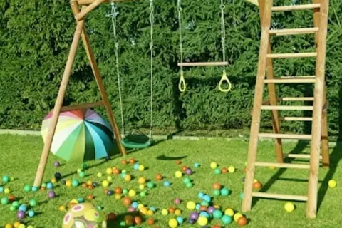 Drewniany plac zabaw ze zjeżdżalnią, huśtawką i drabinką, otoczony kolorowymi piłeczkami i parasolem.