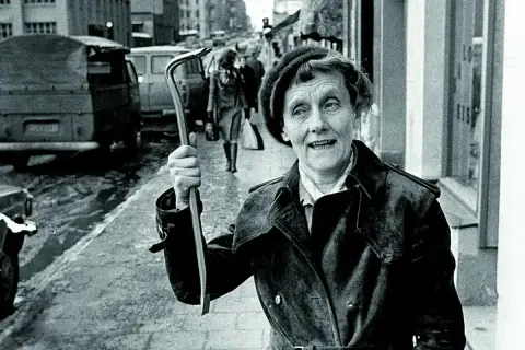 Kobieta z łomem na ulicy, uśmiecha się, jakby mówiła "życie ci niemiłe". W tle samochody i przechodnie.