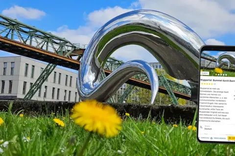 Moderne Skulptur vor Schwebebahn und Gebäude in Wuppertal. Ein Handy zeigt eine Tour durch Barmen, ein tolles Ziel für Ausflugsziele Wuppertal.