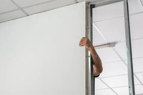 Main d'ouvrier installant une cloison placo avec porte. Le montage de la structure métallique est visible.