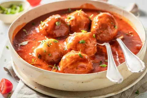 Albóndigas caseras con tomate frito, servidas en un cuenco de cerámica con hierbas frescas y dos cucharas listas para disfrutar.