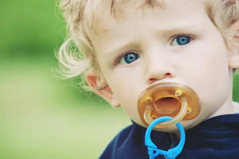 A toddler with blue eyes and blonde hair has a pacifier in their mouth. This image prompts the question: can pacifiers cause cavities?