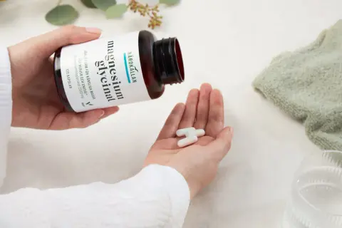 Händer häller upp tre kapslar magnesiumglycinat ur en flaska. Ett uppiggande medicin för nerver och energi.