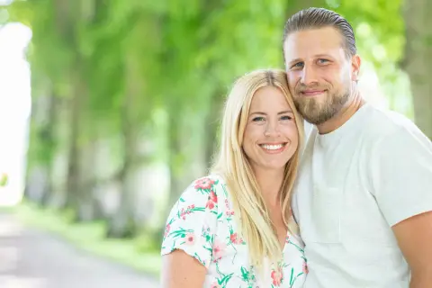 Ett leende par, Nanna Olasdotter Hallberg och hennes partner, njuter av en sommardag utomhus bland gröna träd.
