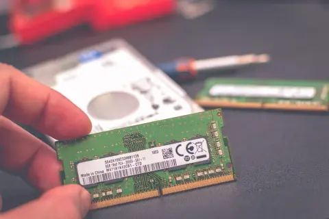 Dłoń trzyma moduł pamięci RAM 8 GB. Obok leży dysk twardy i drugi moduł RAM, sugerując wybór: 8 czy 16 GB RAM w laptopie.