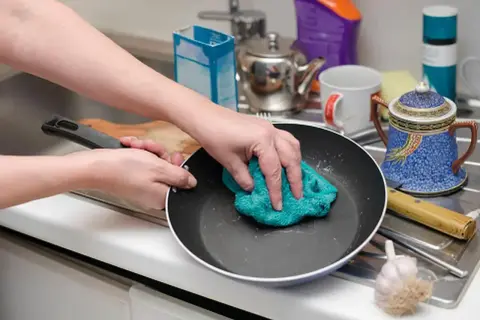 Ręce czyszczą przypalony garnek zieloną ściereczką. W tle zlew, czajniczek i ozdobna cukiernica.