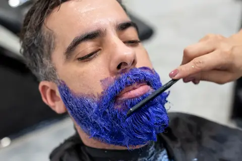 Un homme se fait teindre la barbe en bleu vif par un professionnel. La couleur est appliquée avec une brosse.