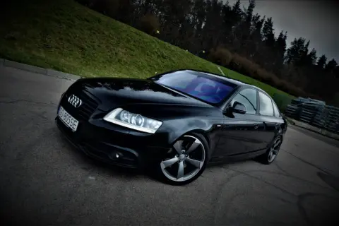 Czarny Audi A6 C6 3.0 TDI Quattro S-line na tle zielonego wzgórza i lasu.