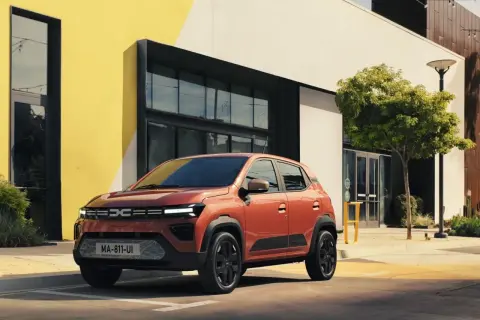 Pomarańczowy Dacia Spring Electric zaparkowany przed nowoczesnym budynkiem z żółtymi akcentami.