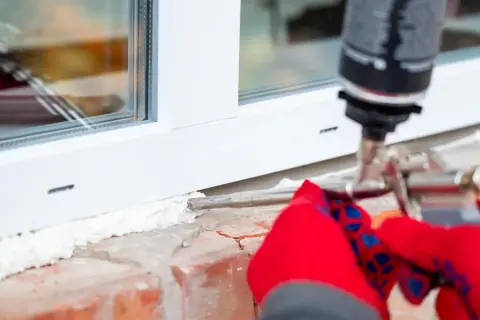 Uszczelnianie okna pianką montażową. Ręce w czerwonych rękawiczkach trzymają pistolet, aplikując piankę na zimny parapet wewnętrzny.
