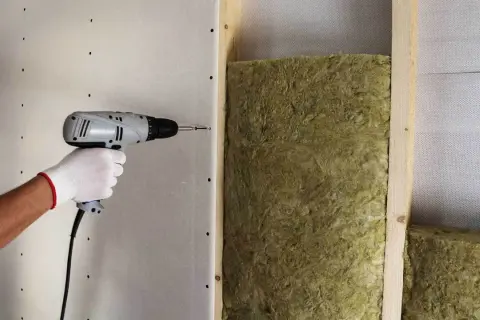 Ręka w białej rękawiczce przykręca płytę gipsowo-kartonową do stelaża, tworząc barierę dla dźwięku. W tle widać wełnę mineralną, kluczową dla wyciszenia pomieszczenia.