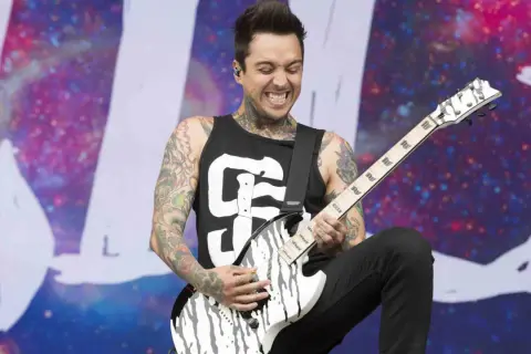 A tattooed guitarist, Tony Pierce the Veil, shreds on a white and black guitar on stage with a cosmic backdrop.