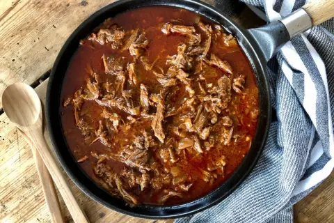 Saftigt pulled beef i en mustig sås i gjutjärnspanna, redo att serveras med träskedar bredvid.