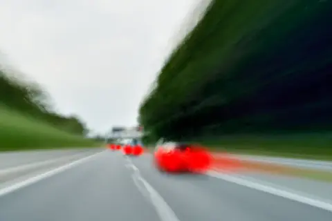 Rozmyte czerwone samochody na drodze, jeden z nich wykonuje wyprzedzanie z prawej strony.