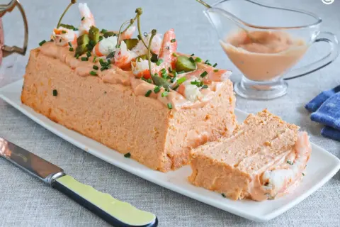 Pastel de pescado decorado con gambas, pepinillos y alcaparras. Una porción ya cortada revela su interior.