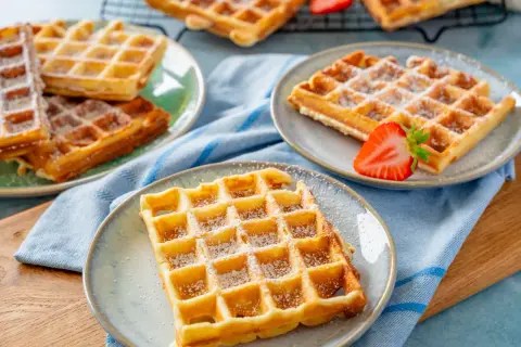 Goldbraune Waffeln, bestäubt mit Puderzucker, warten auf ein leckeres Waffeln Rezept. Eine Erdbeere garniert eine Waffel.
