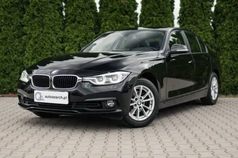 Lśniące czarne BMW 318i stoi na kostce brukowej przed żywopłotem.