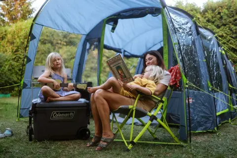 Rodzina odpoczywa pod namiotem podczas festiwalu słońca. Dziecko gra na ukulele, a mama czyta książkę.