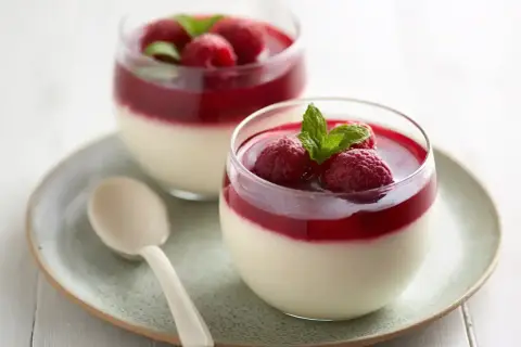 Två glas med krämig pannacotta med hallonspegel, toppad med färska hallon och mynta. En ljus sked ligger på fatet.