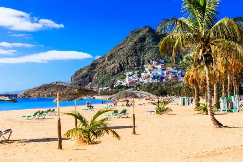 Plage de sable doré, palmiers, parasols et maisons colorées perchées sur une colline. Une invitation à visiter Tenerife pour des vacances de rêve.