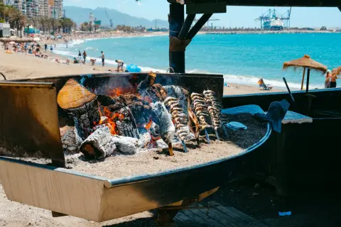 Pescado a la brasa en un chiringuito del paseo marítimo de Málaga. El mar y la playa con gente al fondo.