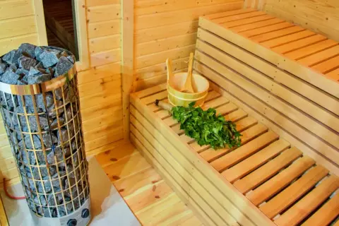 Interno di una sauna fai da te con stufa in pietra, panche in legno, secchiello e frusta di betulla pronti per un relax rigenerante.