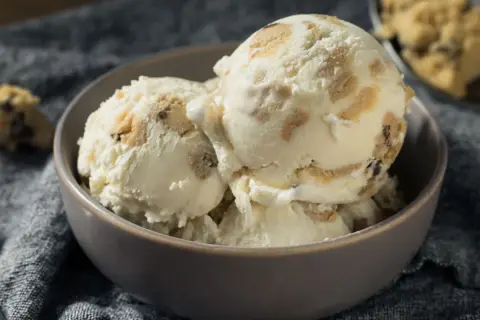 Schüssel mit cremigem Eis, durchzogen von Stückchen aus süßem Keksteig und Schokoladenstückchen. Ein Löffel liegt daneben.