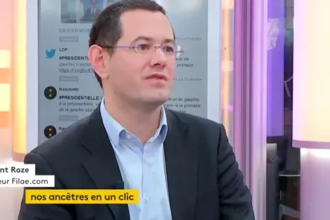 Un homme en costume, lunettes, parle. Le nom de famille gratuit "Filae.com" est visible, suggérant une recherche d'ancêtres.