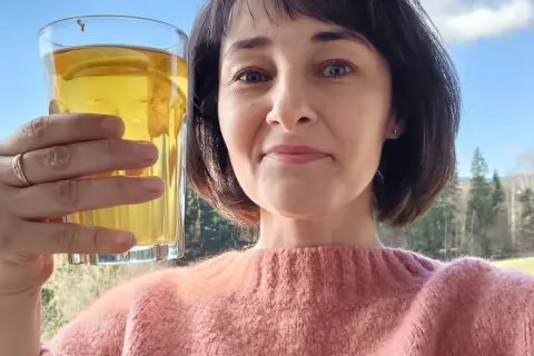 Kobieta trzyma szklankę z napojem, być może to picie pokrzywy, ciesząc się widokiem na las. Efekty zdrowotne napoju są widoczne w jej promiennej cerze.