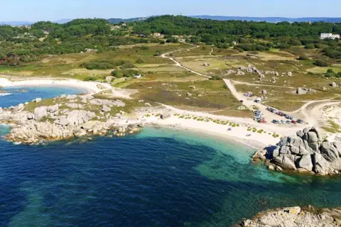 Vista aérea de una playa con arena blanca y aguas turquesas, rodeada de rocas y vegetación. Se ve un aparcamiento con coches cerca de la playa.