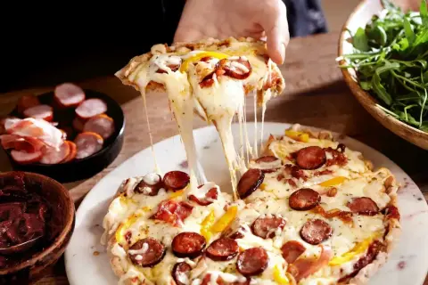 Rozciągający się ser z kawałka pysznej pizza farmerska, z kiełbasą i papryką, obok miseczki z rukolą.