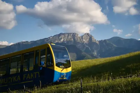 Żółto-niebieski wagonik PKL **wyciąg krzesełkowy na Gubałówkę** wjeżdża na zielone wzgórze, w tle majestatyczne Tatry.